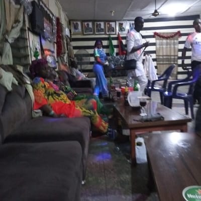 This image depicts an indoor social gathering space in Benin City, Nigeria, characterized by dim lighting. The scene presents multiple individuals, seating arrangements, and beverage-laden tables.
In the foreground, a dark brown upholstered sofa occupies the left side. An adult individual with dark reddish-purple hair, wearing a vibrant multi-colored (orange, yellow, green, black) patterned garment, reclines on this sofa. A light blue wristband is visible on their left wrist. Behind this person, another individual in green attire is partially visible, also seated on a sofa.
In the mid-ground, two dark brown wooden coffee tables are present. The closer table holds a glass with dark red liquid, a white pitcher, several small glasses with beverages, a green bottle, and various containers. A "Heineken" logo is partially visible on the bottom right table surface.
Several individuals are active in the mid-ground and background. An adult female with dark hair, dressed in a light blue top with light green/white sleeves and light blue pants, stands facing away from the camera. To her right, an adult male in a white long-sleeved top and dark trousers walks across the scene, carrying a dark shoulder bag and a folded white cloth in his right hand. Further right, a partially visible adult male wears a white t-shirt with a dark emblem.
The back wall features distinctive horizontal black and white stripes. Above this, at least five framed portraits of individuals are displayed. Red tinsel or decorative lights hang across the top portion of the striped wall, which also features a green and a red triangular banner. A standing fan is positioned against this wall. The left wall displays what appears to be a flat-screen television and additional festive decorations. The floor is dark, with subtle blue light reflections. Dark blue plastic chairs are visible in the mid-ground right. A ceiling fan with an integrated light fixture is also visible overhead.