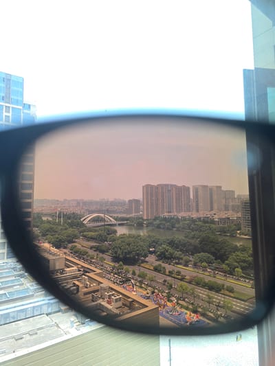 This media file captures an urban landscape viewed through a curved lens, possibly eyeglasses. The scene is dominated by a dense array of modern apartment buildings and office towers, reflecting a bustling city environment. Lush green trees line a wide road that curves through the scene, with a body of water, possibly a river, visible to the left of the buildings. Along the road, numerous parked motorcycles and scooters suggest a common mode of transportation. Several people are visible on the sidewalks and possibly a public area, but their activities are not discernible due to the distance and resolution.
A prominent feature in the mid-ground is a bridge with an arched structure, adding an architectural element to the cityscape. Further along the road, an area with colorful, stylized structures and what appears to be playground equipment or art installations is visible, hinting at public recreational spaces. The sky is hazy, with a reddish-pink hue, suggesting either sunrise or sunset, or perhaps atmospheric pollution. The overall impression is one of a densely populated, modern urban center. The image appears to be taken from an elevated vantage point, likely a building overlooking the city. The lens distortion creates a slightly warped perspective, particularly at the edges of the frame. No discernible text is visible in the image.