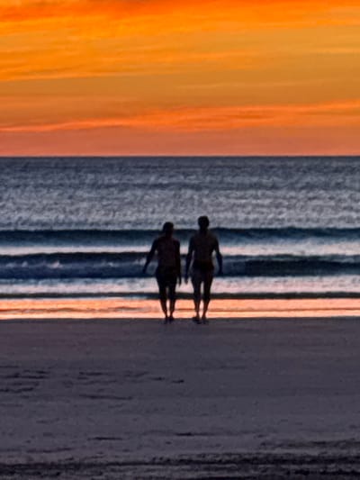 The image is a sideways view of a beach scene at sunset. The sky is visible on the left side of the image and the colours transition from orange to yellow. In the middle of the image is the ocean, represented by horizontal grey lines of varying intensities. On the right side of the image is the beach, which appears to be a dark grey. There are two silhouetted figures on the beach. It is likely the photograph was taken in Cabo Velas, Costa Rica.