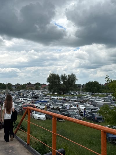 The image depicts a large, outdoor parking lot filled with numerous vehicles, predominantly cars. The parking lot appears to be situated in a semi-rural or suburban area, with trees and some buildings visible in the background, suggesting it might be near the city of Nurkeevo, Russia. The sky is overcast with heavy clouds. In the foreground, a person with long brown hair, wearing a white t-shirt and black pants, is walking away from the camera. The person is carrying a black bag. An orange railing runs across the lower portion of the image, separating the foreground from the parking lot. Several other individuals are scattered throughout the parking lot, some standing near vehicles, and others appearing to be walking. The ground in the parking lot is a mixture of grass and dirt, with some vehicles parked on grassy areas.