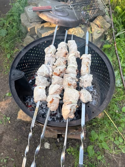 The image shows skewers of raw meat and onions being cooked over hot coals in a metal brazier. The brazier is set on the ground outdoors, surrounded by green grass and some rocks. In the background, there is a metal grill and a lid, suggesting this is a picnic or outdoor cooking setup. The lighting indicates it is daytime, likely outdoors in a natural setting, possibly in a yard or park. The specific location of Sylva, Russia, is not visually evident from the image itself, and can only be inferred if provided as context.