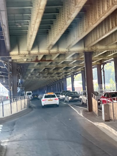 The image shows traffic moving under a large metal bridge structure in the city of Derry/Londonderry, United Kingdom. The bridge appears to be made of steel beams and girders, with a visible underside of latticework and beams. The road below is paved and appears to be a multi-lane route. Several vehicles are visible on the road, including a white car with a yellow sign on its roof, a white van, and several other cars. The environment suggests an urban setting, with the bridge structure dominating the scene. Sunlight is filtering through the gaps in the bridge, creating patterns of light and shadow on the road. There are buildings and trees visible in the distance beyond the bridge.