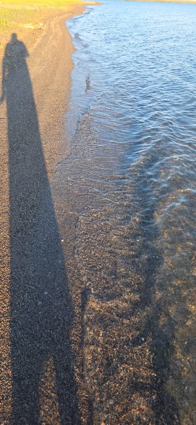 The image depicts a sunlit shoreline on a body of water, likely Issyk-Kul Lake in Kyrgyzstan, given the distinct light and vegetation visible on the upper left. The foreground shows a narrow strip of dark, fine gravel beach. A long, distinct shadow of a person, presumably standing on the beach, stretches across the gravel and into the shallow water. The water itself is clear, revealing the pebbly lakebed beneath the surface. Gentle waves lap at the shore, creating ripples and a shimmering effect on the water. The background is dominated by the tranquil blue expanse of the lake, meeting a pale blue sky at the horizon. The overall impression is one of peaceful solitude and the natural beauty of a lakeside environment.