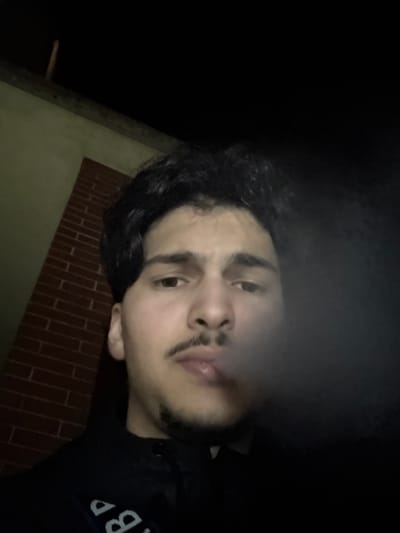 A male individual, appearing as a young adult with dark, wavy hair, a mustache, and chin stubble, is depicted in the image. He is wearing a dark garment with partial light-colored lettering visible on the left side of his chest/shoulder area. A white-gray plume of smoke is emanating from his mouth, partially obscuring the lower facial area. The subject is looking directly at the camera. The background in the upper left quadrant features a light-colored wall positioned above a section of reddish-brown brickwork. A dark wooden beam is visible horizontally above the light-colored wall. The remaining background is uniformly dark, indicating a low-light environment. The scene is located in Holtzheim, France.