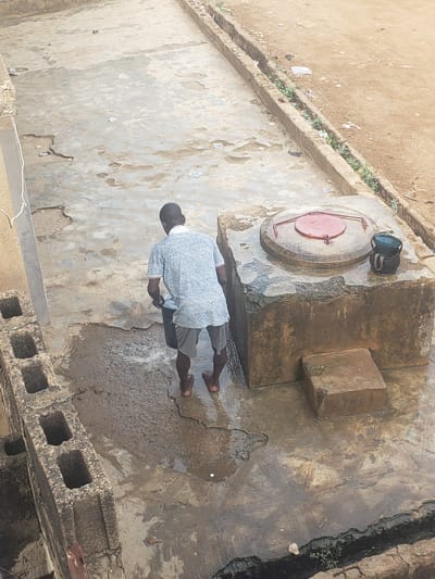 Location: Alalubosa, Nigeria.
Scene: A man stands on a damp, cracked concrete surface. The surface is partially covered with water and debris. The man is facing a circular, metal well cover with a red top. A black bucket rests on the well cover. The man is wearing a light blue shirt, dark shorts, and sandals. He appears to be washing his hands, or something held in his hands. A concrete block wall is partially visible in the lower right corner of the image. A narrow ditch runs along the right edge of the concrete surface. Additional concrete surfaces are seen in the background. There is dirt and some scattered vegetation on the left.
