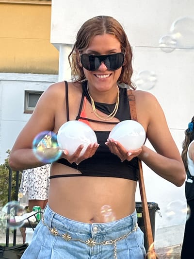 A woman stands outdoors, holding two large white spheres. She is wearing a black crop top, denim shorts, large sunglasses, and a gold chain necklace. Her hair is brown and styled in loose waves. Behind her, a white building and a yellow wall are visible. The environment appears to be a street or a backyard with some greenery. Several bubbles are floating in the air around her. The lighting suggests it is daytime, possibly late afternoon.