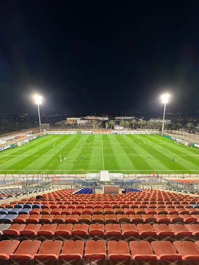 The image captures an elevated view of a football (soccer) stadium in Ness Ziona, Israel, at night. The brightly lit green pitch occupies the central and left portions of the frame, showing clear white markings including the center circle, halfway line, and parts of the penalty areas. Several individuals, likely players or coaching staff, are scattered across the pitch, engaged in what appears to be a training session or warm-up.

To the right, rows of empty stadium seats dominate the foreground. These seats are predominantly a terracotta orange or reddish-brown color, with smaller sections of blue seating visible further down the stands. The stands are devoid of spectators, indicating that this is not a match day.

Two powerful stadium floodlights are prominent features, one in the upper left and another in the lower left, casting strong illumination over the playing field and contrasting sharply with the deep black night sky. Beyond the far side of the pitch, a line of palm trees and various buildings can be seen, with the faint glow of city lights stretching into the distant horizon. The clear, dark sky suggests good weather conditions. No discernible text is visible within the image.