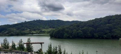 The image depicts a scenic view of a tranquil lake surrounded by lush green hills and dense forests. The sky is overcast with a mix of dark and light clouds, suggesting a potentially cloudy or approaching rainy day. In the foreground, there is a wooden structure, possibly a part of a pier or a gazebo, with some young evergreen trees and flowering plants. A metal fence is visible along the bottom left edge of the frame. The lake's surface is calm with gentle ripples. The hills are covered with a thick canopy of trees, with some buildings peeking through the foliage on the distant slopes. The overall impression is one of natural beauty and serenity, characteristic of a rural or semi-rural landscape. The location is identified as Ikindzhi Nyugedi, Azerbaijan, which implies this is a natural landscape within or near that settlement. There are no people or animals visible in the image.