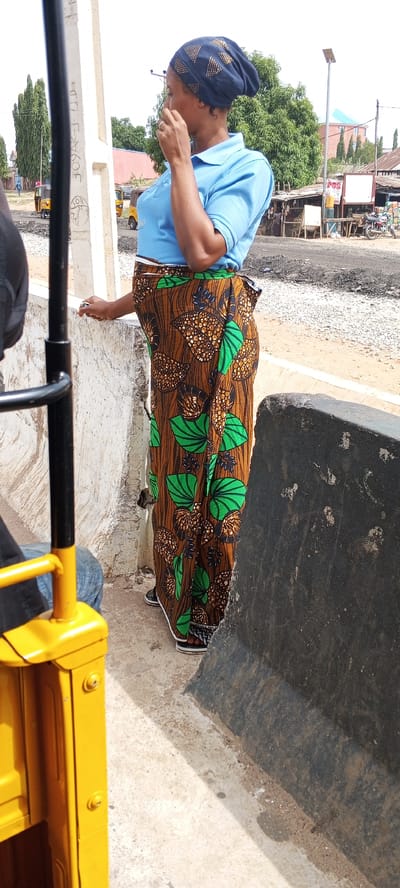 The image, taken in Uke, Nigeria, shows a woman standing on a sidewalk next to a concrete barrier. She is wearing a light blue collared shirt, a brown patterned wrap skirt with green leaf designs, and a dark blue head covering. She is holding her hand to her face, possibly in thought or conversation. In the background, there is a street scene with yellow auto rickshaws ("keke napeps"), buildings, trees, and motorcycles. Some businesses appear to be operating on the side of the road. The road surface appears to be under construction or repair. A portion of a yellow auto rickshaw is visible on the left side of the frame. Another person is seen seated in the foreground left.