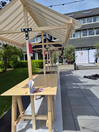 The image shows a temporary outdoor structure with a striped awning, possibly for a market or event. The structure is made of wood and features bolted joints. A wooden table is set up next to the structure, with a book or brochure open on it. Behind the structure is a grassy area and a building with multiple windows, suggesting an urban setting. In the background, a promotional display with the logo "ESCOFFIER LUXEMBOURG" can be seen. The location is likely in Lenningen, Luxembourg, based on the display. There are people visible near the display, but their actions are unclear. The overall scene suggests a daytime event or promotion.