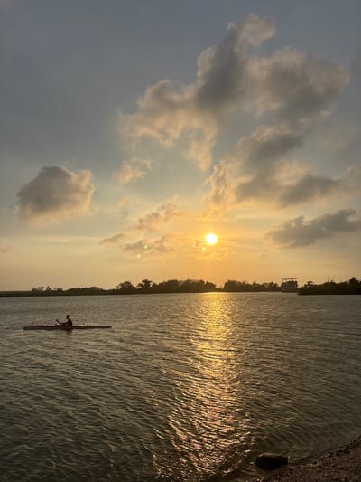 A long shot shows a person rowing in a kayak on a body of water, likely the Chairel Lagoon, at sunset in Tampico, Mexico. The sun is positioned in the center-right of the frame, partially obscured by clouds, casting a golden glow on the water and sky. The water surface is rippled, reflecting the sunlight. A narrow strip of land with trees runs along the horizon, with a small structure visible in the distance. The kayaker is located in the lower-right quadrant of the image.