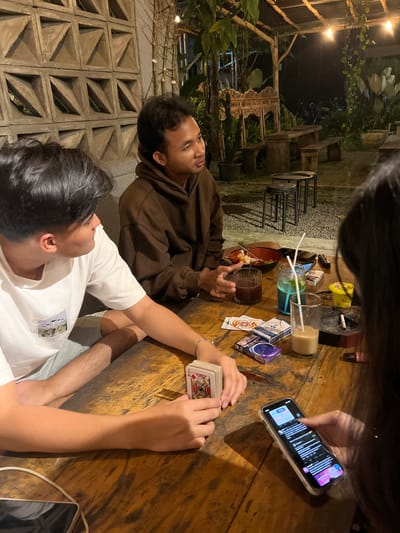 The image depicts a dimly lit outdoor setting, likely a cafe or restaurant, in what is presumably Magelang, Indonesia. Three individuals are partially visible around a wooden table. One person, wearing a brown hoodie, is looking upward, perhaps in conversation. Another, clad in a white t-shirt, holds a deck of playing cards. A third person, partially in view, is engaged with a smartphone.
On the table are various items: a glass of dark liquid, a blue beverage with straws, playing cards (both loose and in boxes), an ashtray with cigarettes, food in a bowl, and lipstick. The table is marked with wear.
Behind the subjects, the scene includes foliage, decorative stonework, and strung lighting, indicating an open-air or semi-enclosed space. The location gives the impression of a casual, evening gathering.