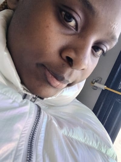 A close-up, angled self-portrait shows a person with dark skin and brown eyes. The person is wearing a light-colored iridescent jacket. The jacket has a visible zipper. The background is blurry and appears to be the interior of a vehicle. The camera angle is tilted, with the person's face angled towards the viewer. The image quality is slightly grainy.