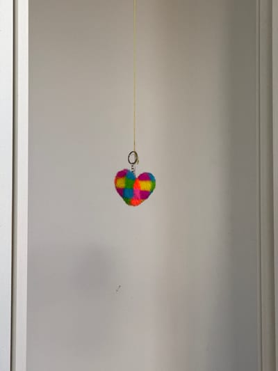 A fluffy, heart-shaped keychain with alternating squares of bright colors like yellow, green, blue, and pink, hangs from a thin yellow string against a neutral-toned wall. The keychain is attached to a silver ring. The wall appears to be indoors, with subtle shadows and texture. The scene is framed by vertical elements on the left and right sides, suggesting it might be within a doorway or a similar architectural feature. There are no people or animals visible in the image, and no specific landmarks or context to place it in Brasília, Brazil.