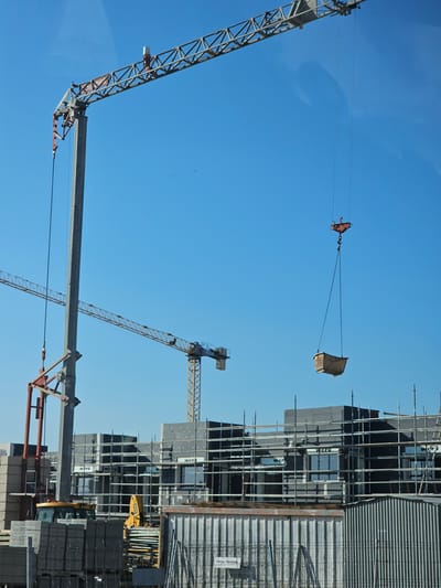 This image depicts a construction site in Cuarte de Huerva, Spain, under a clear blue sky. Two tower cranes are prominently featured. The larger crane on the left has a significant boom extending upwards and to the right. A smaller, red-colored attachment is visible near the base of its vertical support. A concrete box is suspended in the air from the larger crane by cables. In the middle ground, a yellow construction vehicle, possibly a backhoe loader, is partially visible. The construction site itself shows building structures in various stages of completion, characterized by dark grey exterior panels and scaffolding. Several windows with white frames are present on the buildings, some bearing the "WERU" logo. In the foreground, stacks of building materials, including what appear to be concrete blocks and thin planks, are organized. A corrugated metal structure is situated in the lower right portion of the frame. The overall scene suggests active construction work.