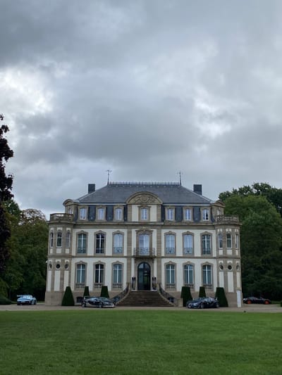 The image displays Château Saint-Jean, the Bugatti headquarters, located in Dorlisheim, France, under an overcast sky. The multi-story, classically-styled mansion features a light-colored facade with darker stone accents, a mansard roof, and a central grand staircase leading to the main entrance. Dormer windows and decorative balconies are visible across the upper stories. A well-maintained green lawn occupies the foreground, leading to a semi-circular gravel driveway in front of the building. Three high-performance automobiles are parked on the gravel drive: a light blue/silver sports car with a prominent rear wing is positioned on the left, a dark grey Bugatti supercar is situated centrally directly before the staircase, and a dark grey Bugatti supercar with orange wheel accents is partially visible on the right. Lush green trees flank the building in the background. No individuals are present in the scene.