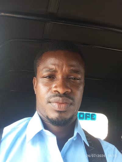 A man is taking a selfie inside what appears to be the cabin of a vehicle. He is wearing a light blue collared shirt. The background is dark, with a bright white light and some text that appears to say "OFE" visible in the upper right. A date stamp in the lower right corner reads "26/07/2026 14:15". The overall setting suggests a casual, possibly on-the-go moment. There is no specific information in the image to identify the city of Alagada, Nigeria.