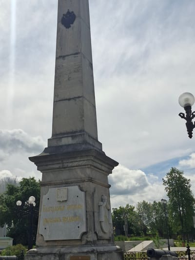 This media file captures a tilted, low-angle view of an obelisk monument with an inscription. The monument itself is made of stone, with a tall, tapering shaft that has a dark, possibly soot or stain, mark on its left side. The base of the obelisk features decorative architectural elements and a prominent stone plaque with golden Cyrillic text. The inscription on the plaque reads: "БЛАГОДАРНЫЕ ПОТОМКИ ХРАНИТЬ ПОДВИГОВЫ".
To the right of the obelisk, a cannon barrel is visible, suggesting a historical or military context. Beyond the monument, a wrought-iron fence encloses a grassy area with trees and lampposts. In the background, modern architecture with a distinctive striped pattern can be seen peeking through the trees. The sky is filled with large, white, puffy clouds, indicating a bright, possibly daytime setting. A decorative streetlamp with a glass globe is also visible at the top of the frame.
The scene appears to be a public space, likely a park or square, commemorating a historical event or individuals. The presence of the obelisk, cannon, and inscription points to a memorial or monument. The weather seems fair, with a mix of sun and clouds. The orientation of the shot, from a low angle looking upwards, emphasizes the grandeur and height of the monument. The location context provided is Kungur, Russia.