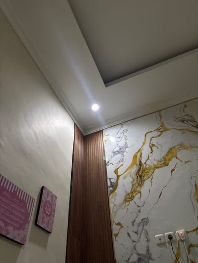 This indoor shot features an angled view upwards, showcasing an interior space with a combination of wall and ceiling decor. The top right area of the frame exhibits a wall with a white marble-like pattern infused with gold accents and bordered by a strip of wooden paneling. An electrical outlet is installed on the marble wall.

To the left, a section of the ceiling appears with a white border. A bright light source, possibly from a bulb, casts a starburst effect.

In the lower portion of the image, two decorative, rectangular plaques are visible on what appears to be a light-colored wall. They are adorned with floral patterns and Arabic text.

Given the religious inscriptions and the common use of Arabic script in Islamic contexts, the location is likely in an area with a significant Muslim population. Binong is a region in Indonesia with a Muslim majority, making it a plausible location. No individuals are visible, and the scene is static.