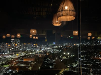 The image presents a night-time panoramic view of the illuminated cityscape of Floridablanca, Colombia, observed from an elevated indoor vantage point, likely a high-rise building or a rooftop establishment. The foreground is dominated by a series of suspended pendant lamps, featuring woven, basket-like shades that emit a warm, amber light. Two larger, conical lamps are prominently positioned in the upper right quadrant, with several smaller, similarly styled lamps distributed across the top and mid-sections of the frame. Reflections of these interior lights and the building's internal structure are visible on a large vertical glass window or panel that separates the indoor space from the exterior view.
Beyond the glass, the sprawling urban environment of Floridablanca extends into the distance under a dark night sky. Numerous multi-story buildings, characterized by their illuminated windows, form dense clusters of light across the landscape. Veins of light suggest roads and highways with active vehicle traffic. Darker, unlit areas within the city likely represent parks, unpopulated zones, or low-rise structures. In the far background, the indistinct silhouettes of hills or mountains are discernible against the horizon. No individuals are visible, nor are specific actions or interactions discernible within the static scene.