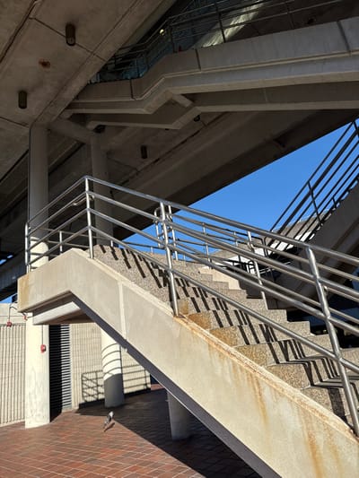 An architectural shot, looking upward, featuring concrete structures, metal railings, and a glimpse of blue sky. Concrete supports and beams form sharp angles against the sky, while a metal staircase with railings cuts diagonally across the frame. The composition emphasizes the interplay of lines and shadows, and the contrasting textures of concrete and metal. The shot's orientation creates a sense of disorientation and emphasizes the height of the structures.