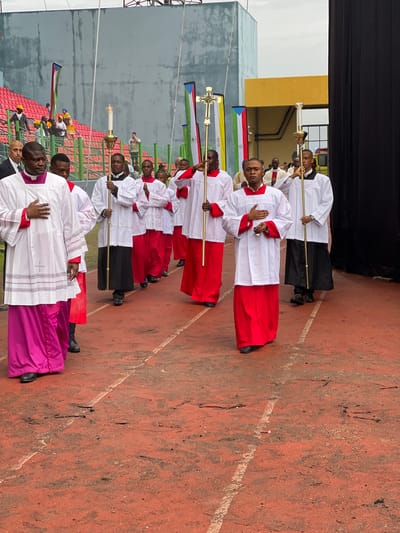 A procession of approximately ten men in religious vestments stands in a line outdoors on a reddish-brown paved surface, possibly an athletic track, indicated by a faint white line on the ground. The first person in the line, on the left, wears a purple cassock with a white surplice and holds a crozier, suggesting a bishop or high-ranking clergy. The subsequent individuals wear white surplices over either red or black cassocks. Many of them hold tall staffs topped with golden crosses or liturgical candles. One person near the front holds a thurible (incense burner).

The event appears to be a religious ceremony or procession. The subjects have solemn expressions, and their posture is formal, with some holding their hands clasped or placed on their chest.

In the background, there are various structures, including a yellow building and grey, perhaps industrial, buildings under an overcast sky. Behind the participants, rows of red stadium-style seating are visible, suggesting the event is taking place in an arena or large public space. Colorful banners or flags are also visible in the background; one distinctly features the design of the South Sudanese flag (black, red, green, blue triangle, white stripes). The overall lighting is bright and even, characteristic of daylight on an overcast day. No discernible text is visible on any objects or attire.
