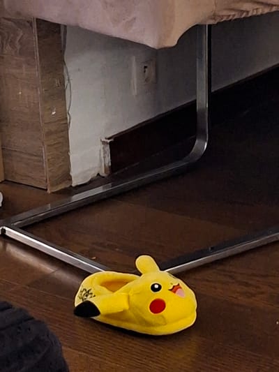 The image shows a close-up view of a yellow plush slipper shaped like the character Pikachu from the Pokemon franchise. The slipper is lying on a wooden floor. To the left of the slipper, there is a partial view of a beige upholstered object and a dark wooden surface. The background consists of a wooden floor and a light-colored wall with some exposed wooden planks at the bottom. A metal railing or edge runs vertically along the right side of the image, separating the slipper from the wall. The lighting appears to be from an indoor source, suggesting the image was taken inside a home or building. There is no visible text in the image. The scene appears to be a casual, domestic setting in Evere, Belgium.