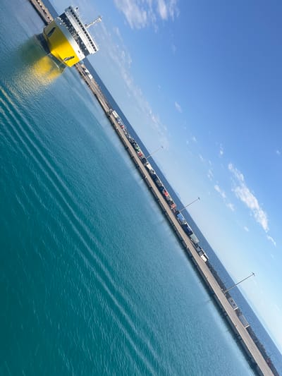 A bright yellow and white ferry is docked at a port in Kastro-Kyllini, Greece. The ferry, which appears to be a car ferry, is positioned at the head of a concrete pier. The pier extends into the turquoise water, lined with numerous trucks and what appear to be shipping containers. The sky above is a clear blue with scattered white clouds. The angle of the photograph is tilted, giving a dynamic perspective of the scene. The water exhibits a pattern of ripples, indicating calm conditions. The sun is shining, casting reflections on the water and illuminating the side of the ferry.