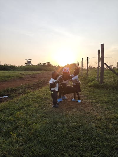 The image depicts an outdoor scene at sunset or sunrise in Ang'orom ward, Kenya. Three individuals are centrally positioned: an adult female crouching, flanked by two young children. The adult female, wearing a light-colored sleeveless top, dark trousers, light blue slide sandals, and a red and white patterned head covering, smiles faintly towards the viewer while interacting with the children. To her left, a young child stands, dressed in a white t-shirt, dark trousers, and dark shoes, with one arm extended. To her right, another young child stands, wearing a light-colored long-sleeved shirt, dark skirt or shorts, and light blue slide sandals, appearing to lean into the adult.

The ground in the immediate foreground and around the subjects is covered in green grass. A dirt path is visible extending from the middle-left towards the background. In the distant left background, a multi-tiered metal tower structure, featuring multiple platforms and what appear to be tanks or equipment, stands amidst sparse vegetation under a clear horizon. To the right of the subjects, a fence line constructed with vertical wooden posts and horizontal wires stretches into the distance. The sun is low on the horizon, positioned behind the individuals, casting an orange and yellow glow across the sky and illuminating the scene from the rear. The upper sky transitions to a pale blue and white.