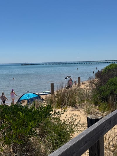 This media file captures a sunny day at a beach or bay, with the perspective slightly rotated, showing a clear blue sky, calm water, and a sandy shore with vegetation.

In the background, a long, wooden pier or jetty extends far into the tranquil, light blue-green water, lined with numerous vertical posts. Several small figures are visible in the distance, both on and near the pier, and in the water near the shore further away.

Closer to the viewer, the sandy shore is lined with green bushes and dry grass. On the sand, near the water's edge, a person is seen relaxing under a black umbrella. Further to the left, in the shallow water, another individual is wading.

In the foreground, two or more people are standing near a striking blue and black inflatable object, possibly a paddleboard or a small sail, resting on the sand or just at the water's edge. One person appears to be wearing a purple hat. A wooden beam or railing, possibly part of an elevated walkway or viewing platform, frames the upper right portion of the image, suggesting the photo was taken from a slight elevation.

The scene depicts a leisurely day with people enjoying the water and the shore under a bright, clear sky, indicating pleasant weather. There is no visible text in the image. The setting suggests a calm coastal area, consistent with the context of Frankston, Australia.