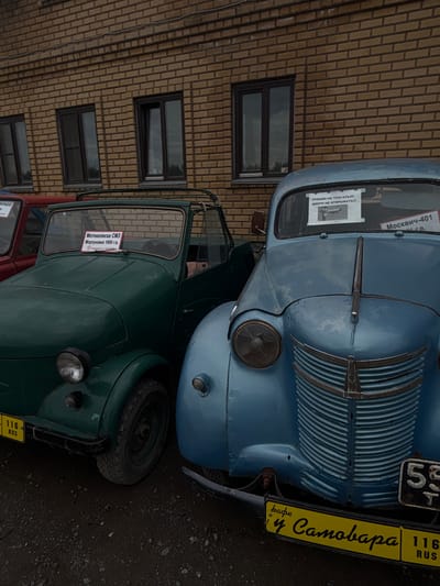 This image shows two vintage cars parked in front of a brick building. The car on the left is green and appears to be a Motokolesa SMZ. It has a sign on the windshield that reads "Мотоколяска СМЗ Моргуновка 1959 г.в." and "сделано в СССР". The car on the right is light blue and appears to be a Moskvich-401. It has a sign on its windshield that reads "РУКАМИ НЕ ТРОГАТЬ!!!! ДВЕРИ НЕ ОТКРЫВАТЬ!!!!" and "Москвич-401". Below the Moskvich, a yellow license plate with "у Самоварa" is visible, along with a smaller plate showing "116 RUS". The background is a textured brick wall with three windows. The ground is a dark, gravelly surface. The scene is static and captures a moment of display or exhibition of these classic vehicles. No people are visible in the image. The location is identified as Bol'shoi Mashlyak, Russia.