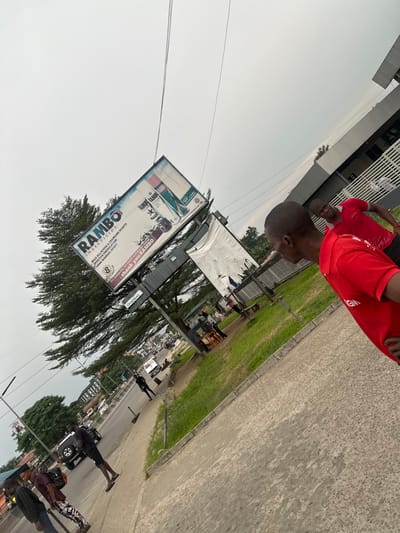 The image showcases a Rambo insecticide billboard that reads "Thank You Rambo." The advertisement features two cartoon characters, Lambi and Lambo Rage, along with details about the insecticide's formulation and its effectiveness against flying and crawling insects. The word "Global" is printed at the bottom right corner. Two people in red shirts are standing in the background.