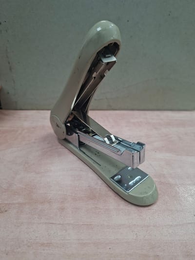 A cream-colored stapler rests on a light pink wooden surface. The stapler is open, revealing its metallic inner workings and a row of staples. The background is a textured, grey wall. The image is taken from a slightly elevated angle, focusing on the stapler. There are no people or animals present in the scene. The environment appears to be indoors, possibly an office or workspace.