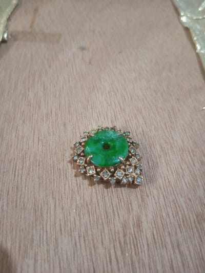 An ornate piece of jewelry is positioned in the lower-right quadrant of a light brown wooden surface. The object features a central, circular, translucent green stone, approximately 1.5 cm in diameter, with a small central perforation. This green stone is secured by four prongs within a gold-toned metal setting. Surrounding the central stone are numerous smaller, clear, faceted stones, each set in a square-shaped bezel, forming an asymmetrical, embellished border around the main stone. The wooden surface exhibits distinct vertical grain patterns and minor surface imperfections. The location of the scene is Chandragiri, Nepal.