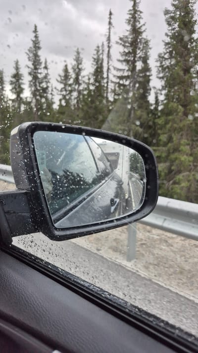 The image shows a close-up view of a car's side mirror during a rainy day, reflecting the surrounding environment. The mirror itself is speckled with raindrops, and the housing is dark gray. In the reflection, the side of another vehicle, possibly a truck or trailer, is visible, as well as a road and a guardrail. Behind the road, a dense forest of evergreen trees is present under a cloudy sky. The interior of the car, specifically the door panel, is visible in the foreground, also showing raindrops on the window. The location is identified as Nesland, Norway, suggesting a driving scene on a roadway in a forested area.
