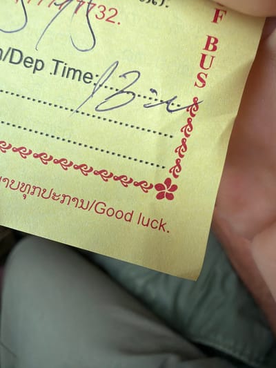The image shows a close-up of a yellow ticket. The ticket has red text on it, including the word "BUS" vertically aligned on the right side. There is also a handwritten entry next to "Dep Time:" that appears to be a signature or time. The bottom of the ticket has a decorative border with a red flower and text in what appears to be Lao script followed by "Good luck." The ticket is held by a person, with a hand and finger visible on the right side. The background is blurry and appears to be fabric, possibly clothing. There is no indication of the specific location within the city of Phakhao, Laos.