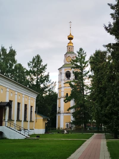 This image depicts a scene in the city of Vasil'ki, Russia, featuring a prominent yellow and white bell tower with a golden dome and cross, set against a cloudy sky. To the left of the bell tower is a similar yellow and white building, possibly a church or monastery building, with a staircase leading up to its entrance. The area is surrounded by lush green grass and tall trees, some of which are coniferous and appear to be partially obscuring the bell tower. A paved walkway, with red and gray bricks, runs diagonally through the foreground. Two individuals are visible sitting on benches in the background, one to the left and another further back on the right, appearing to be enjoying the park-like setting. The overall impression is one of a peaceful, historical religious site.