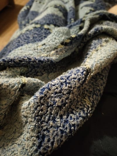 This is a close-up, abstract image of a textured blue and white fabric. The fabric appears to be a blanket or a similar soft textile, with a raised, looped texture. The pattern consists of broad stripes of light blue and white, interspersed with darker blue areas. The texture creates a rough, almost fuzzy appearance. The lighting is dim, suggesting an indoor setting or a time of day when natural light is low. The background is dark and out of focus, providing no discernible environmental cues. There is no visible text or identifiable subjects such as people. The image focuses solely on the tactile and visual qualities of the fabric itself.