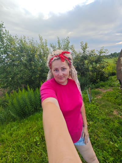 A selfie is taken outdoors in Novoe Mart'yanovo, Russia, on a cloudy day. The subject, a woman with blonde dreadlocks and a red bandana, is wearing a pink t-shirt and denim shorts. She is standing in a grassy area with trees and bushes behind her. There is a wooden object with writing on it to the right of the frame. Two wooden stakes are visible in the grass to the left. The woman is holding the camera with her right arm extended, capturing her upper body and a portion of the surrounding environment.