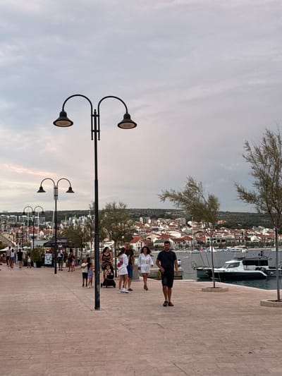 The image depicts a promenade in the city of Primošten, Croatia. The promenade is paved with light-colored bricks. In the foreground, two lampposts with double lamps are visible. Several people are walking along the promenade. On the left, a group of people including children are walking, and a woman with a baby in a stroller is also present. Towards the right, a man is walking towards the camera, and a woman is walking away. In the background, a body of water with several boats is visible, and beyond that, the city of Primošten is spread out with its characteristic red-tiled roofs and buildings. The sky is cloudy.