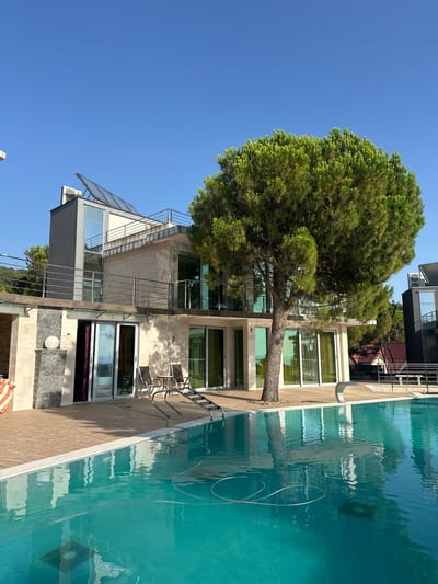 The image shows a modern villa with a swimming pool in Brca, Montenegro. The villa has multiple levels, with large glass windows and balconies. Solar panels are visible on the roof. A large pine tree is situated in the foreground, casting a shadow over the pool area. Two chairs and a table are placed on the wooden deck next to the pool. The swimming pool is filled with clear turquoise water, and a pool cleaning hose is visible. In the background, other buildings and trees can be seen. The sky is clear and blue.