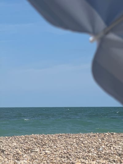 The image depicts a beach scene with a clear blue sky above the ocean. In the foreground, a pebbled beach is visible, with various shades of grey and tan stones. The ocean is a deep blue-green with gentle waves, and the horizon line is sharply defined. In the upper right portion of the image, a portion of a large, blue and white striped umbrella is blurred and out of focus, casting a shadow that obscures part of the sky. The image was captured on a sunny day. The location is the beach of Vinaròs, Spain.