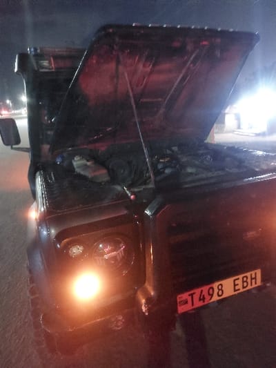 The scene depicts a dark-colored utility vehicle, likely a Land Rover Defender type, parked on a paved surface at night in Msata, Tanzania. The vehicle's hood is fully open, revealing components within the engine bay. The front left headlamp and an auxiliary light are illuminated, casting an orange glow forward and onto the ground. The vehicle's front license plate, "T498 EBH," is clearly visible. Bright artificial lights from the background indicate proximity to a road or urban area. No individuals are visible in the frame. The overall environment is dark, illuminated primarily by the vehicle's lights and distant artificial sources.