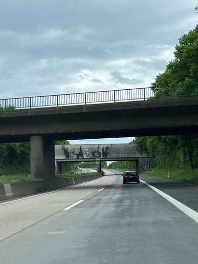 The image depicts a black car traveling on a wet, gray asphalt road in Bergheim, Germany. The road is marked with white lines. The scene is viewed from a moving vehicle. An overpass bridges the road; it's constructed of gray concrete with a black metal railing. Graffiti is visible on the overpass's support columns. Green trees and vegetation line the sides of the road. The sky is overcast with gray clouds.