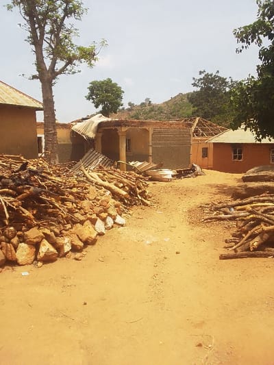 The image shows a scene of destruction in a residential area of Kwal, Nigeria (latitude: 9.807, longitude: 8.658). The roofs of two mud-brick houses have been damaged and are missing sections. Debris, including wood and metal, lies scattered across the ground. A pile of rocks and wood sits in the foreground. Trees and vegetation are visible in the background. There are no people present. The damage appears to be caused by a strong windstorm or other natural disaster.