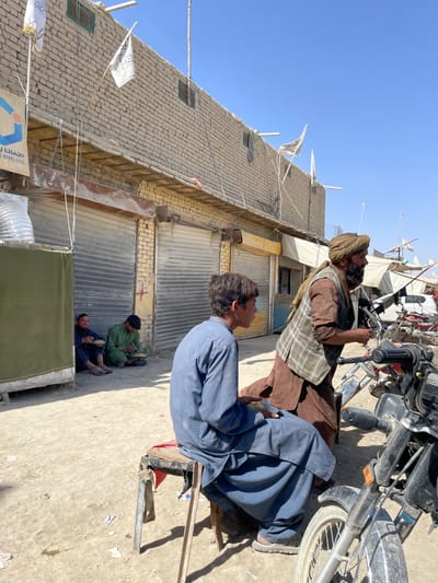 The image depicts an outdoor street scene in Spin Buldak, Afghanistan, under a clear, bright blue sky. A low-rise brick building with multiple closed metal roll-down shutters, indicative of shops, forms the background on the left. Several white flags bearing black Arabic script are affixed to poles along the building's facade and other makeshift structures. A partially visible blue and white sign displaying "Wireless" is present on the far left.
In the foreground, a young male with dark hair wears a loose blue shalwar kameez and sits on a makeshift wooden stool with a red fabric top, facing right while holding a small, light-colored rectangular object in his lap. To his right, an older male with a dark beard and mustache wears a brown turban, a brown shalwar kameez, and a plaid vest. He stands next to a parked motorcycle, leaning slightly forward, appearing to interact with it or an unseen object. The handlebars and front wheel of a black motorcycle are prominent on the right side of the frame.
In the mid-ground, to the left, two other males are seated on the dirt ground in the shade of a green fabric partition. One male wears dark blue attire, and the other wears a green shalwar kameez. Both are actively eating from yellow bowls. The ground is unpaved, consisting of packed dirt with scattered debris. Further down the street on the right, additional makeshift shelters or stalls with white coverings are visible, alongside other parked motorcycles and a white vehicle.