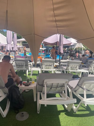 The image depicts a poolside area, likely at a resort or club, situated in Zazymia, Ukraine. In the foreground, a man with dark hair and a goatee is seated on a lounge chair, wearing dark swim trunks. To his right, a small white table holds two glasses with drinks, a container with food, and a red cup. A black backpack rests on the artificial green turf in front of the man.
Numerous white plastic lounge chairs with grey cushions are arranged throughout the scene, some occupied and others empty. In the middle ground, a swimming pool is visible, with several people enjoying the water. A woman in a patterned bikini stands by the pool, facing away from the camera. Another woman in a bikini walks past the pool. Children can be seen playing in and around the pool.
Several large, light-colored umbrellas provide shade over parts of the seating area. In the background, trees and buildings are partially visible, suggesting an outdoor recreational facility. The overall atmosphere appears relaxed and summery, with people engaged in various leisure activities.
