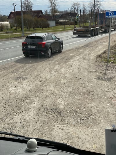 A black HAVAL car, license plate O367OO47, is parked on the right side of a two-lane road. Across the road, a flatbed truck with a blue "70 РОСАВТОДОР" sign is traveling. The area appears to be Bol'shoe Teshkovo, Russia, based on the license plate region. The environment includes sparse trees and structures that appear to be buildings and a traffic warning sign.