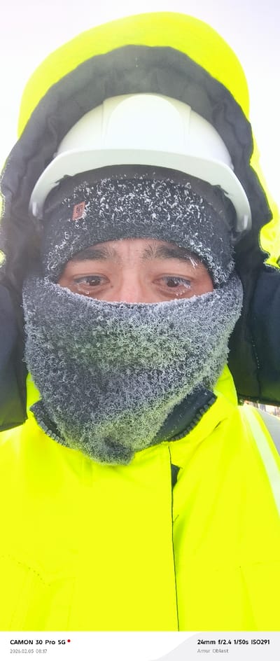 A close-up shows a person wearing a white hard hat and a bright yellow, hooded winter coat. The person's face is partially covered by a gray, fleece-like neck warmer, heavily frosted with ice. Ice is also visible on their eyebrows and eyelashes. The photograph was taken with a CAMON 30 Pro 5G, 24mm lens at f/2.4, 1/50s, ISO291 in Amur Oblast on February 5, 2026, at 08:17.