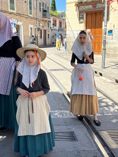 Two young girls in traditional Mallorcan dress stand on a cobblestone street in Sóller, Spain. The first girl wears a brown-patterned skirt, a white apron, and a patterned head covering and plays with a red ball. The second girl wears a green skirt, a white apron, and a straw hat over a patterned head covering. The street has tram rails. The background features buildings typical of Sóller. A pharmacy sign, "Apotecaria Tortell," is visible on one of the buildings. In the distance, more people dressed in similar traditional attire can be seen on the street.